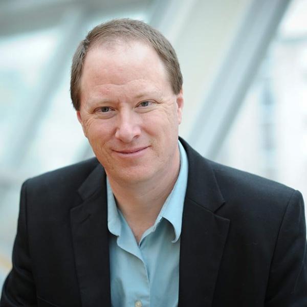 Professional photo of a man wearing a light blue button-up with a black blazer, sitting on an outdoor staircase in the city. 